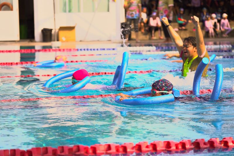 Young swimmers racing with foam pool noodles while a coach cheers in the water