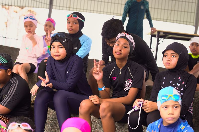 Swimmers in caps and goggles sitting together, some making peace signs, waiting for their race